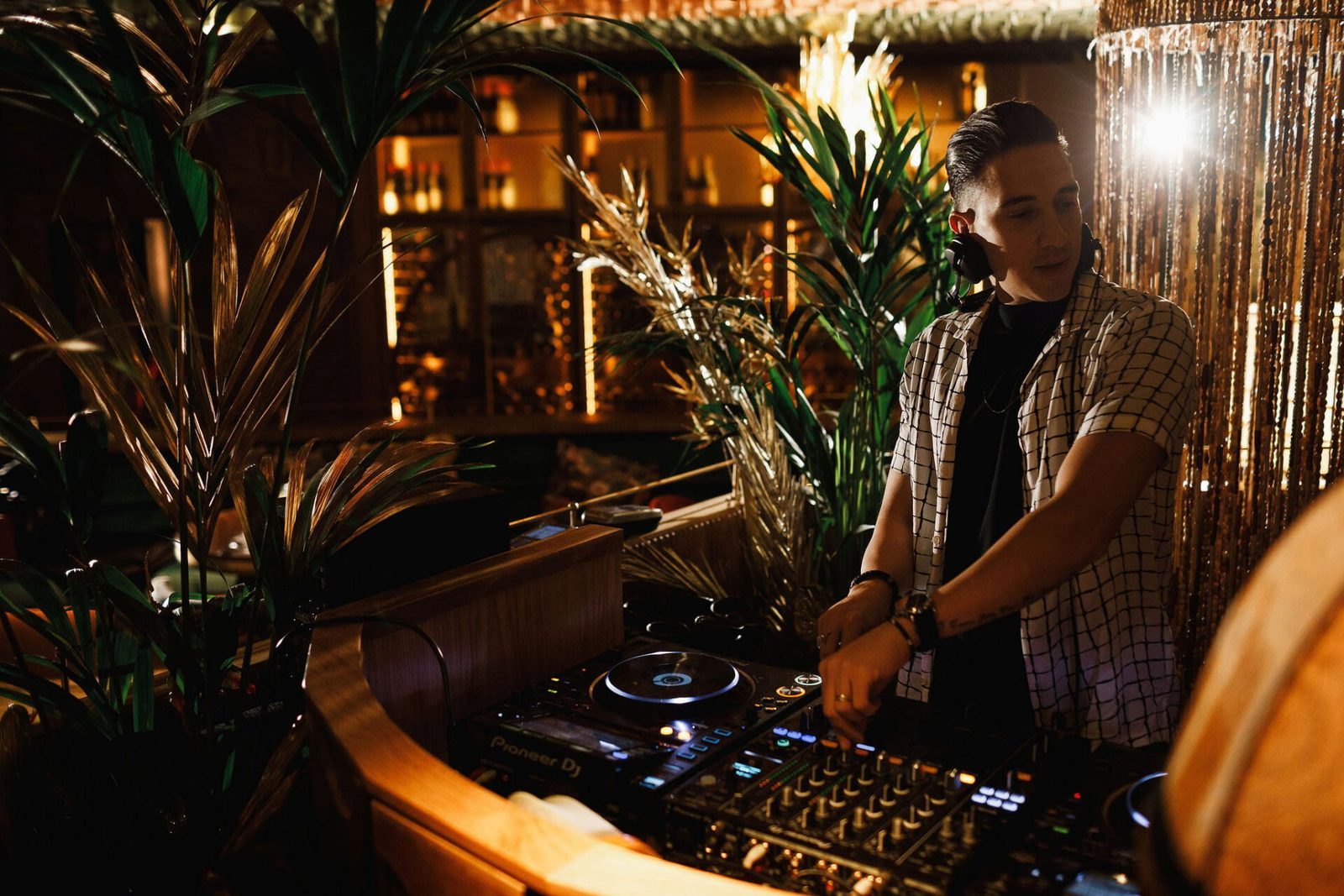 A DJ stands behind a wooden booth surrounded by lush tropical plants, adjusting controls on a mixer and turntables. Warm, golden lighting illuminates the cozy, upscale bar area behind him, casting a soft glow over bottles and decor. The atmosphere feels intimate and vibrant, blending music with a tropical lounge ambiance