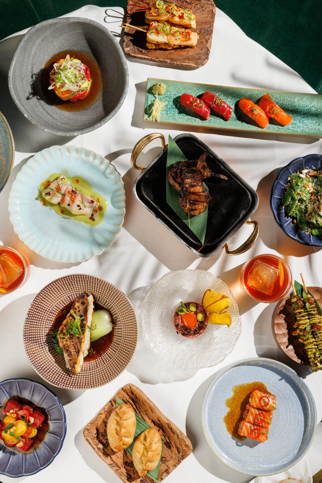An abundant overhead spread of Amazónico's set lunch dishes, including sushi, grilled fish, tartare, bao buns, and cocktails, served on eclectic artisan plates and boards.